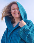 Keltainen designin petroolinsävyiseen vohvelikylpytakkiin pukeutunut nainen hymyilee tuulessa ja auringossa kylpytakin hupun takaa.