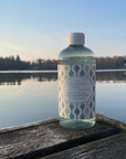 Kaurilan saunan Biohajoava siivousaine 500 ml muovipullossa laiturilla. Taustalla tyyni järvi ja vastaranta matalata paistavan auringon hämyisessä valossa.