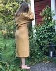 Saunatonttu Mailin Pellavakylpytakkiin pukeutunut nainen seisomassa avojaloin Kaurilan saunan pihalla.