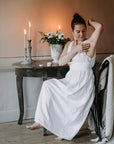 Valkoiseen yömekkoon pukeutunut nainen levittämässä Kaurilan Saunan vegaanista deovoidetta kainaloihinsa, antiikkisen puupöydän äärellä. Taustalla tunnelmallisesti palavia mehiläisvahakynttilöitä ja valkoisia kukkia maljakossa.