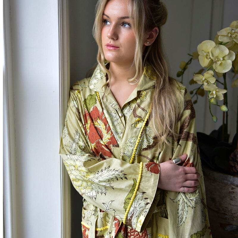 Vaaleahiuksinen nainen katselee ikkunasta ulos Powell Craftin heleän oliivinvihreä puuvilla-aamutakki yllään. 