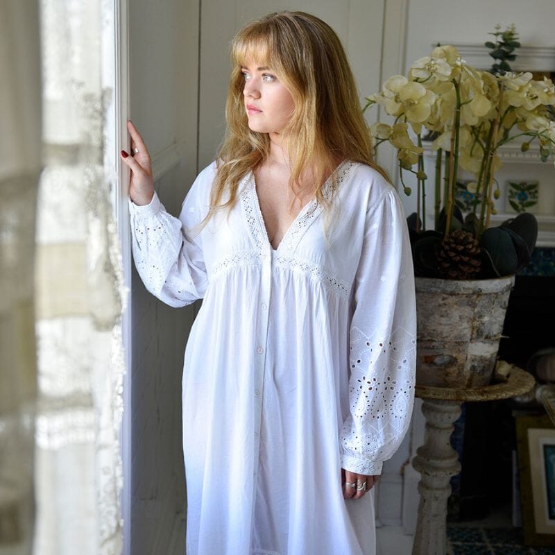 Vaaleahiuksinen nainen seisoo pitsiverhoisen ikkunan edessä ja katselee siitä ulos, yllään Powell Craftin Clarissa-yömekko.