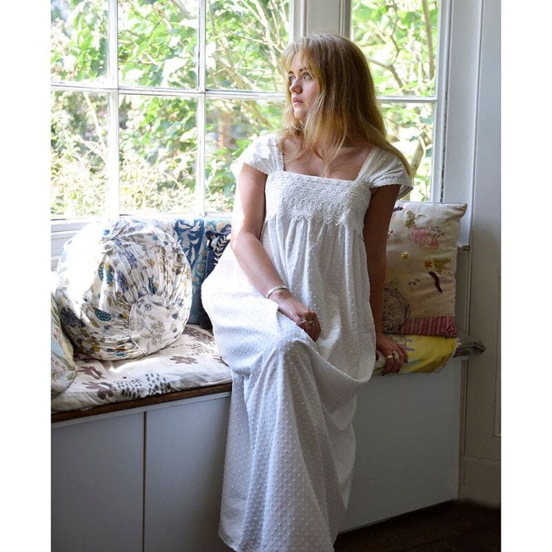 Nuori nainen istuu mietteliäänä vanhan talon ikkunasyvennyksessä, hempeiden tyynyjen koristammalla penkillä. Naisen yllä on Powell craftin valkoinen Maud-yömekko.