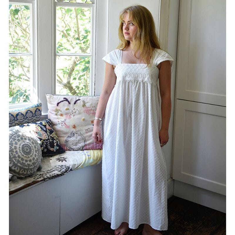 Powell Craftin Swiss dot -puuvillainen Maud-yöpaita ikkunan edessä seisovan naisen yllä. Ikkunan alla penkillä on värikkäitä tyynyjä ja ikkunan takana kesäinen puu.