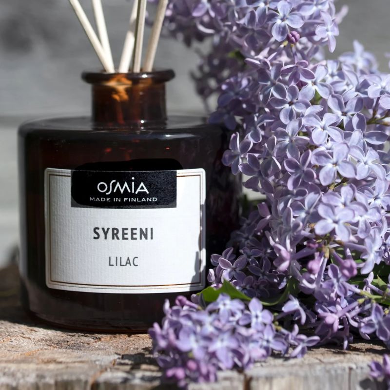 Matala lasinen ruskea pullo auringonvalossa. Osmian syreeni -huonetuoksu on asetettu kannon päälle ja sen vieressä köllöttää kauniin sävyisiä liiloja syreenin kukkia. 