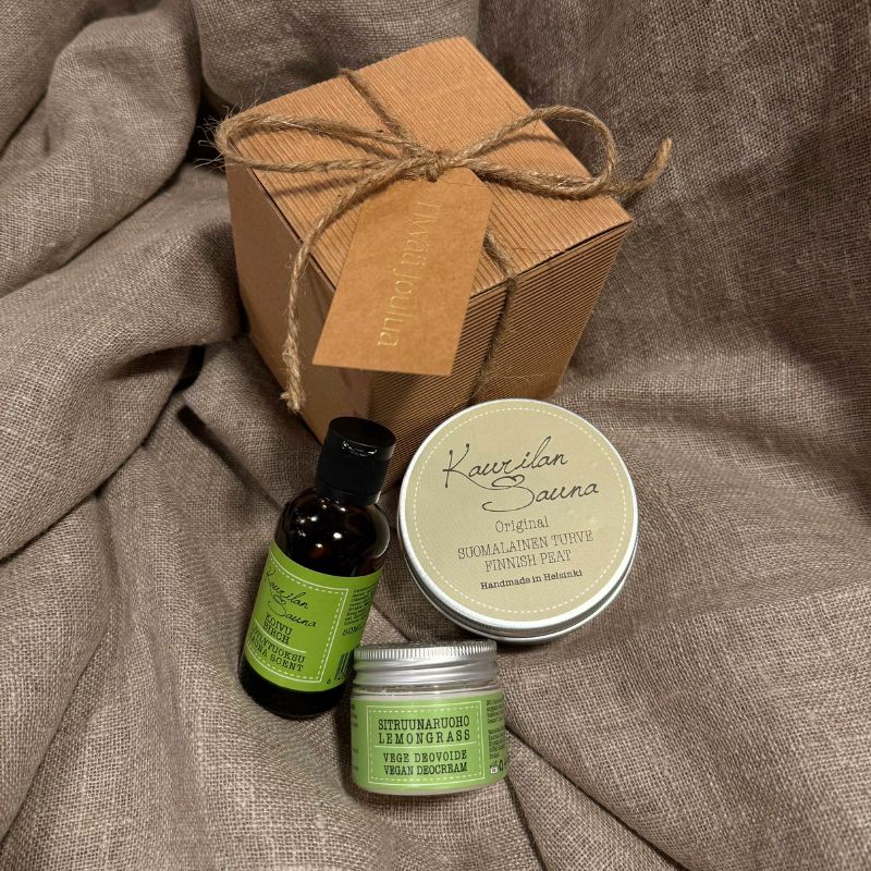 Kaurilan Saunan Tontun Saunasetti Ⅰ - Lahjapaketti vaaleanruskean kankaan päälle aseteltuna. Lahjapaketti on tyylikästä luonnollisen ruskean sävyistä aaltokartonkia ja se on sidottu juuttinarulla, johon on kiinnitetty samaa sävyä oleva pakettikortti, jossa lukee kultaisella 'Hyvää joulua'. Paketin vieressä on siihen kuuluvat vihreäetikettiset Koivu-löyöytuoksu, Suomalainen turvenaamio ja Vegaaninen sitruunaruoho-deovoide.