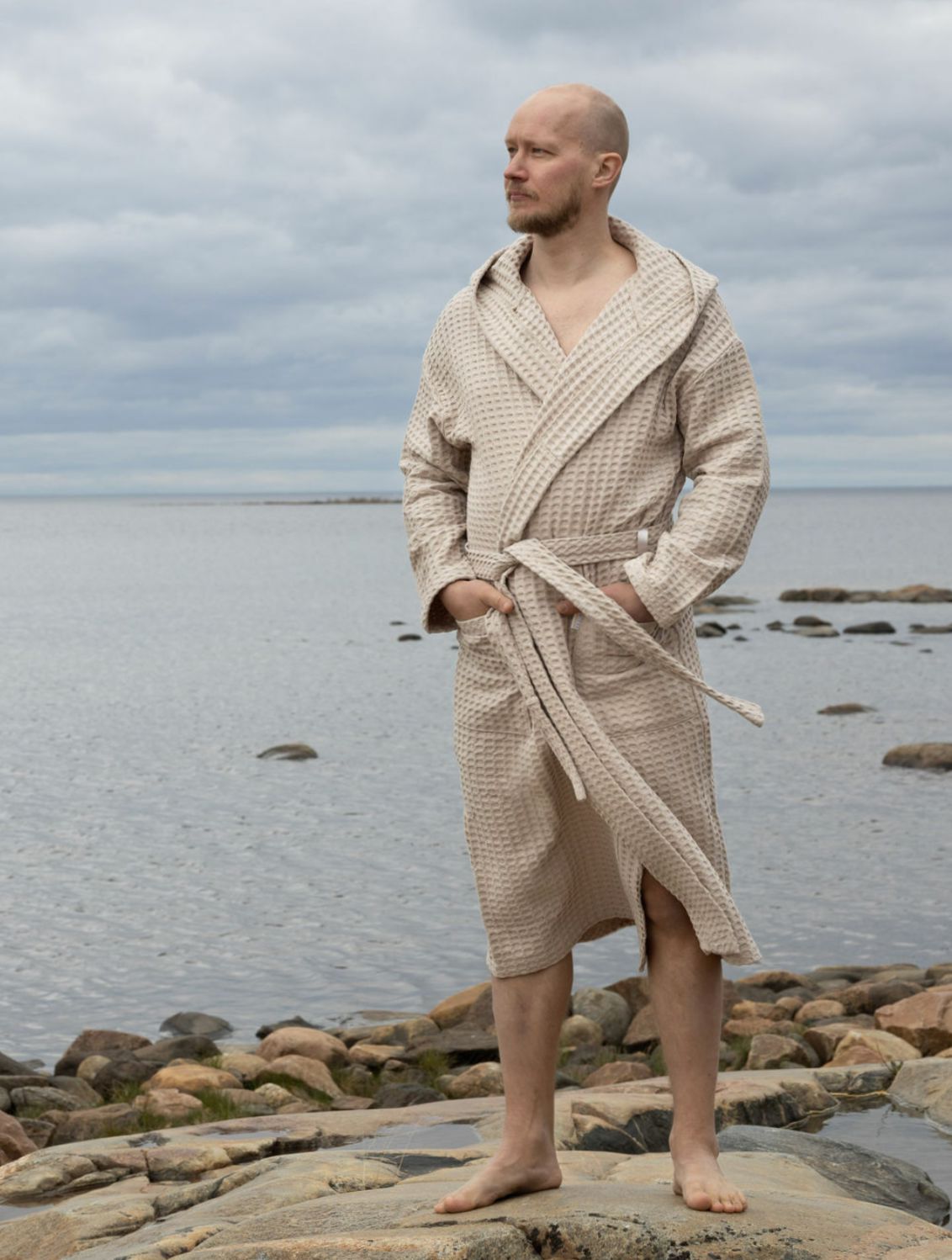 Hiekansävyiseen Keltainen designin vohvelikylpytakkiin pukeutunut mies tuijottelee merenrannalla kaukaisuuteen.