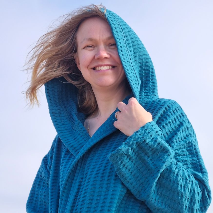 Keltainen designin petroolinsävyiseen vohvelikylpytakkiin pukeutunut nainen hymyilee tuulessa ja auringossa kylpytakin hupun takaa.