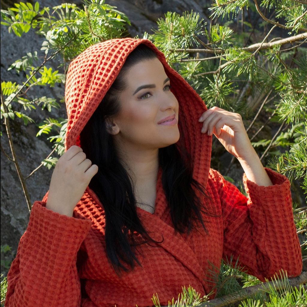 Tiilenpunaiseen Keltainen designin vohvelikylpytakkiin pukeutunut nainen huppu päässään hymyilee aurinkoisessa metsässä.