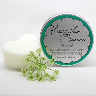 Kaurilan saunan hajusteeton hoitoainepala ja sen metallirasia valkoisella taustalla, etualalla kukkia.