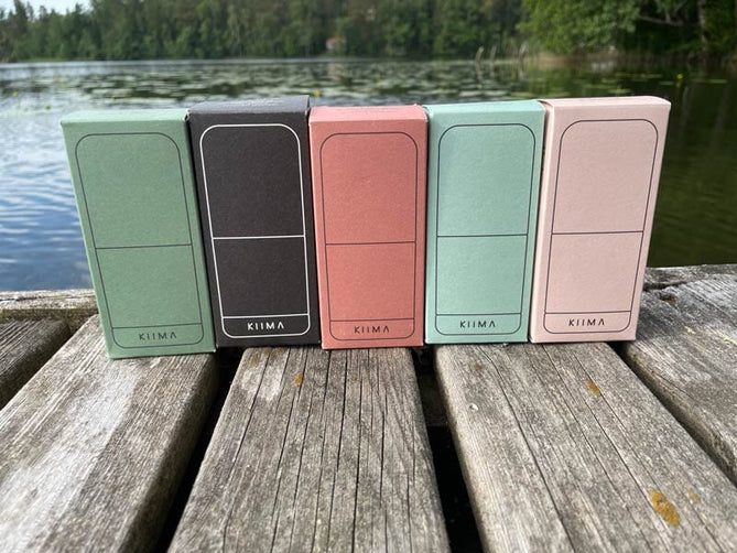 KIIMA -deodoranttiaplikaattorit laiturilla paketeissaan, sävyissä salvia, hiilii, roosa, minttu ja puuteri. Taustalla vettä, lumpeenlehtiä ja metsää. 