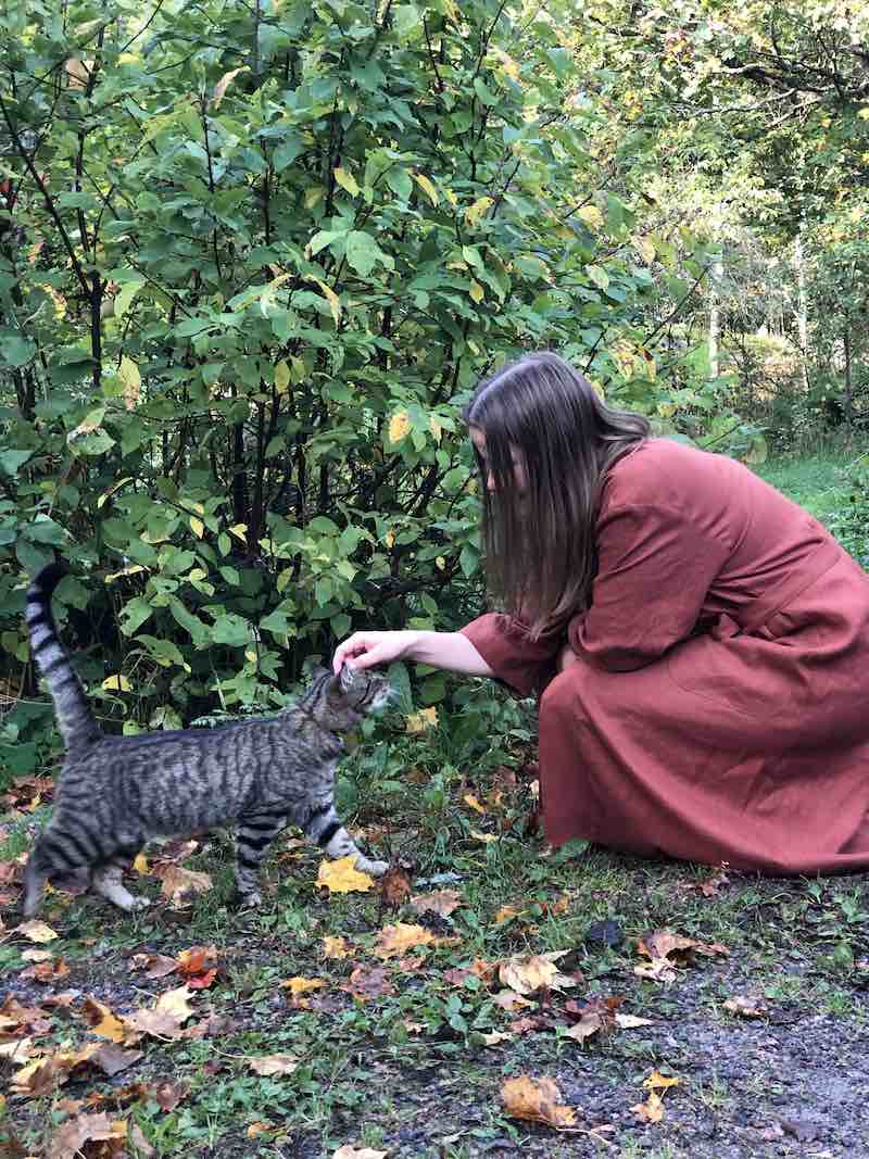 Tummahiuksinen nainen rapsuttelee harmaaraitaista kissaa syksyisellä pihamaalla, päällään Kaurilan saunan ruosteenvärinen Saunatonttu Mailin pellavakylpytakki.