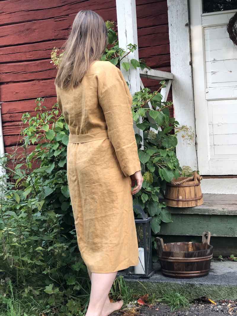 Saunatonttu Mailin sinapinvärinseen pellavakylpytakkiin pukeutunut nainen Kaurilan Saunan ovella.