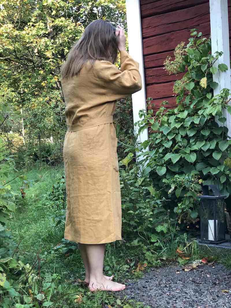 Saunatonttu Mailin Pellavakylpytakkiin pukeutunut nainen seisomassa avojaloin Kaurilan saunan pihalla.