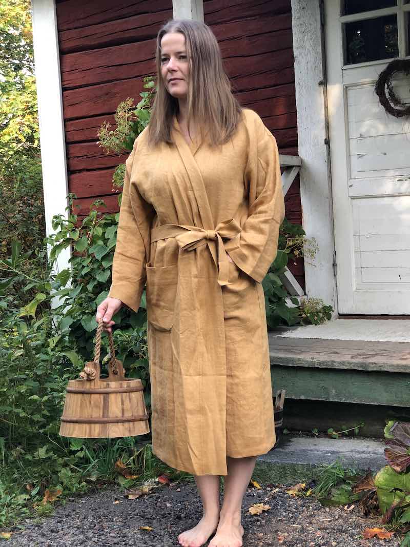 Sinapinväriseen Saunatonttu Mailin Pellavakylpytakkiin pukeutunut nainen seisomassa Kaurilan Saunan ovella puinen kiulu kädessään.