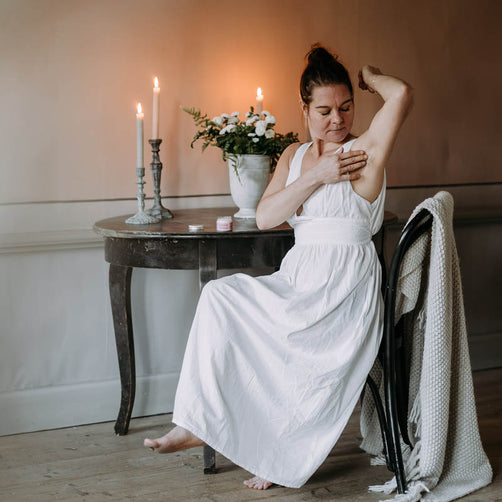 Valkoiseen yömekkoon pukeutunut nainen levittämässä Kaurilan Saunan vegaanista deovoidetta kainaloihinsa, antiikkisen puupöydän äärellä. Taustalla tunnelmallisesti palavia mehiläisvahakynttilöitä ja valkoisia kukkia maljakossa.