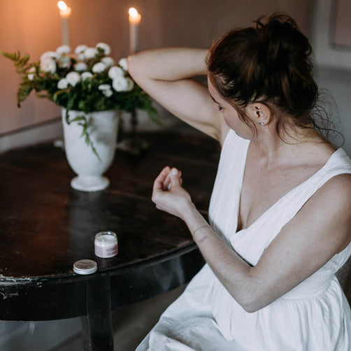 Valkoiseen yömekkoon pukeutunut nainen levittämässä Kaurilan Saunan vegaanista deovoidetta kainaloihinsa tumman antiikkisen pöydän ääressä. Taustalla käsintehtyjä mehiläisvahakynttilöitä ja valkoisia kukkia maljakossa. 