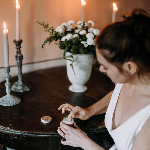 Valkoiseen yömekkoon pukeutunut nainen annostelemassa Kaurilan Saunan vegaanista deovoidetta antiikkisella puupöylällä lepäävästä purnukasta. Taustalla kauniisti palavia mehiläisvahakynttilöitä ja valkoisia kukkia maljakossa.